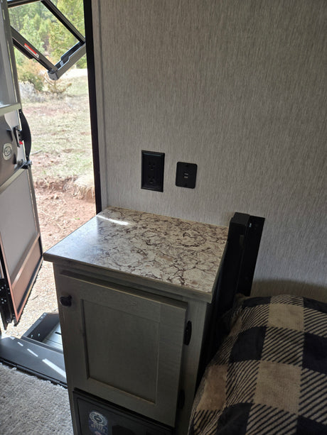 RV rental interior: nightstand with power outlets, open door to trees, and a cozy plaid blanket on the bed.