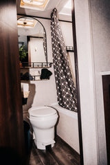 RV rental bathroom featuring a toilet, compact shower with patterned curtain, mirror, and built-in storage.