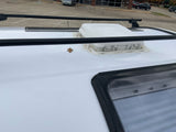 Roof of a white RV rental with a fan vent, skylight, and black roof racks. A fallen leaf rests nearby.