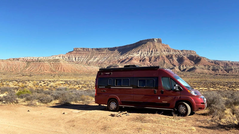 A red Travato RV rental camper van parked on dirt in front of vast red rock mountains — photo 1