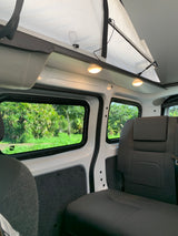 Cozy RV rental interior: pop-top roof, comfortable seating, and large windows revealing lush green nature outside. Ideal for travel.