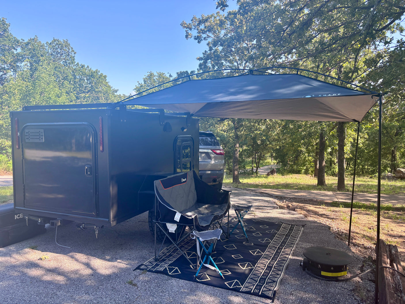 This RV rental features a black travel trailer with a spacious canopy, comfy chairs, and a grill, perfect for a relaxing outdoor setup among the trees.
