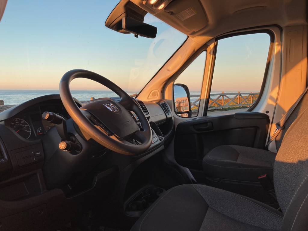 Enjoy your RV rental road trip! Inside a spacious van, the driver's area is visible. Golden light fills the cabin, overlooking an ocean sunset.