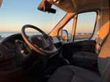 Enjoy your RV rental road trip! Inside a spacious van, the driver's area is visible. Golden light fills the cabin, overlooking an ocean sunset.