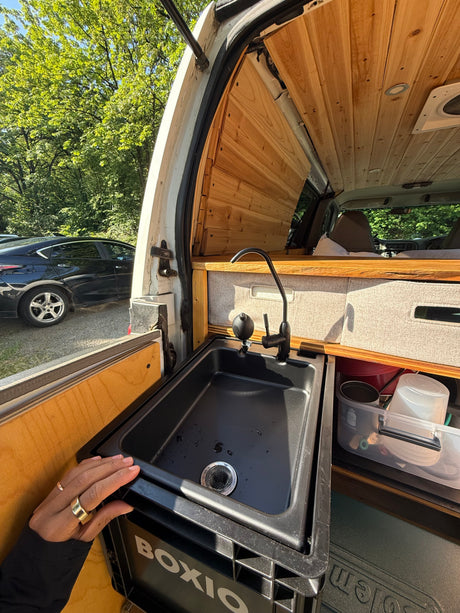 Inside an rv rental, a black sink with a modern faucet and wood paneling. Organized storage and a glimpse of nature outside.