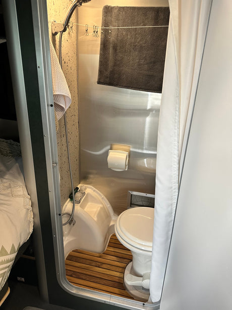 RV rental compact wet bath: integrated toilet, shower, towel line with clips, and wooden floor mat for efficiency.