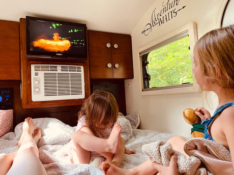 Inside an RV rental, two kids unwind on a soft bed. One watches TV, the other looks out the window at green trees. Relaxation on wheels!