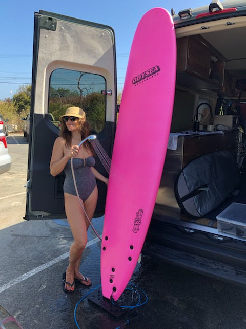 Woman uses an RV rental's outdoor shower to rinse off herself and her pink surfboard after a surf.