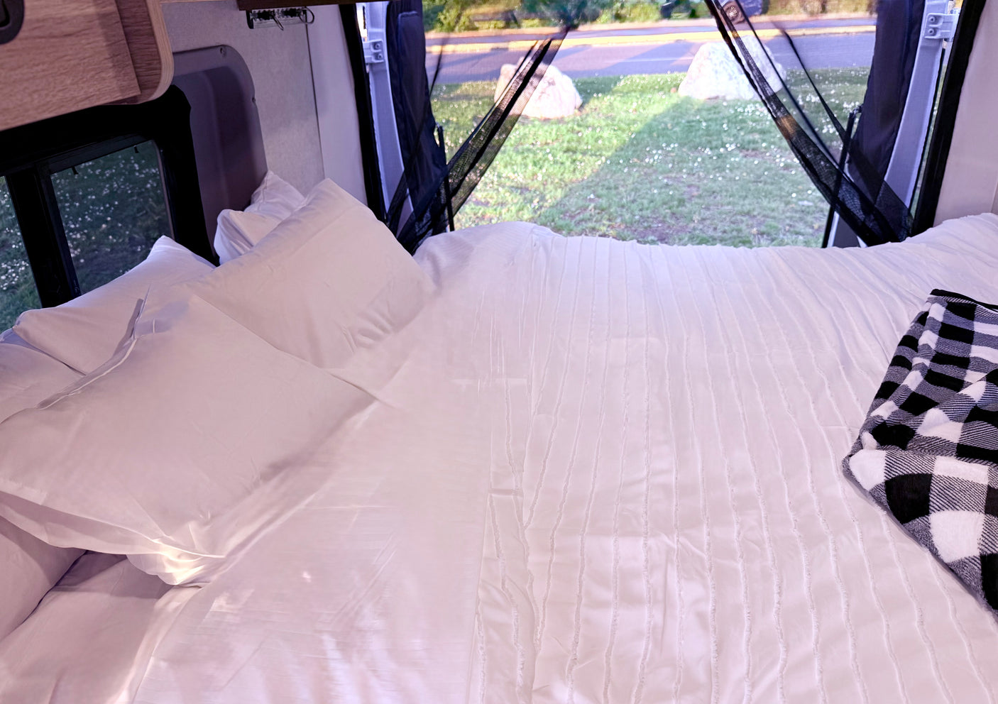 Cozy bed in an RV rental with white sheets, a buffalo check blanket, and a view of nature through screened windows.