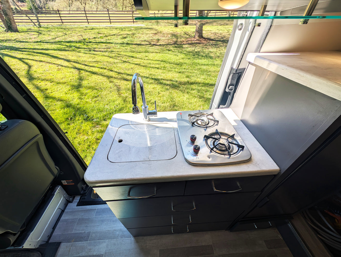 **RV rental** kitchenette: Inside a camper van, a compact kitchen with sink, faucet, and two-burner stove overlooks a sunny field.