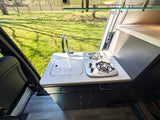 **RV rental** kitchenette: Inside a camper van, a compact kitchen with sink, faucet, and two-burner stove overlooks a sunny field.