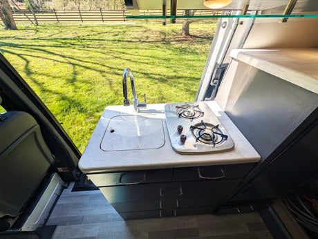 **RV rental** kitchenette: Inside a camper van, a compact kitchen with sink, faucet, and two-burner stove overlooks a sunny field.