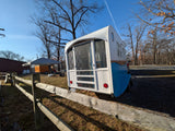 Unique blue & white custom RV rental, resembling a tiny home, parked by a wooden fence under a clear sky.