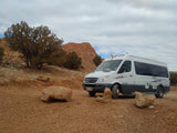 White Roadtrek Sprinter RV rental parked on a cracked red dirt road, amidst desert trees and red rock formations.