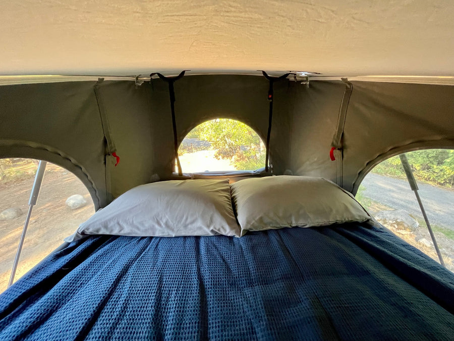 RV rental rooftop tent interior: cozy bed with blue blanket, light pillows, and nature views. Perfect for camping!