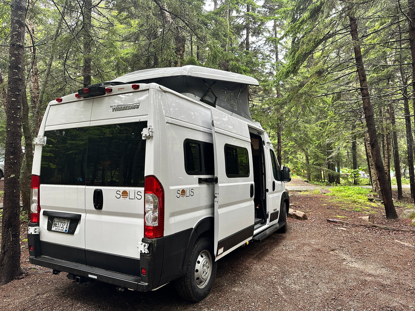 White Winnebago Solis rv rental, pop-top open, parked on a forest path. Ideal for nature travel.