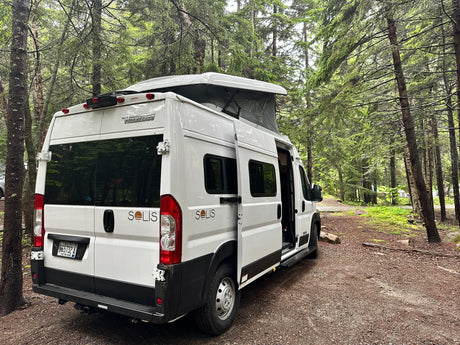 White Winnebago Solis rv rental, pop-top open, parked on a forest path. Ideal for nature travel.