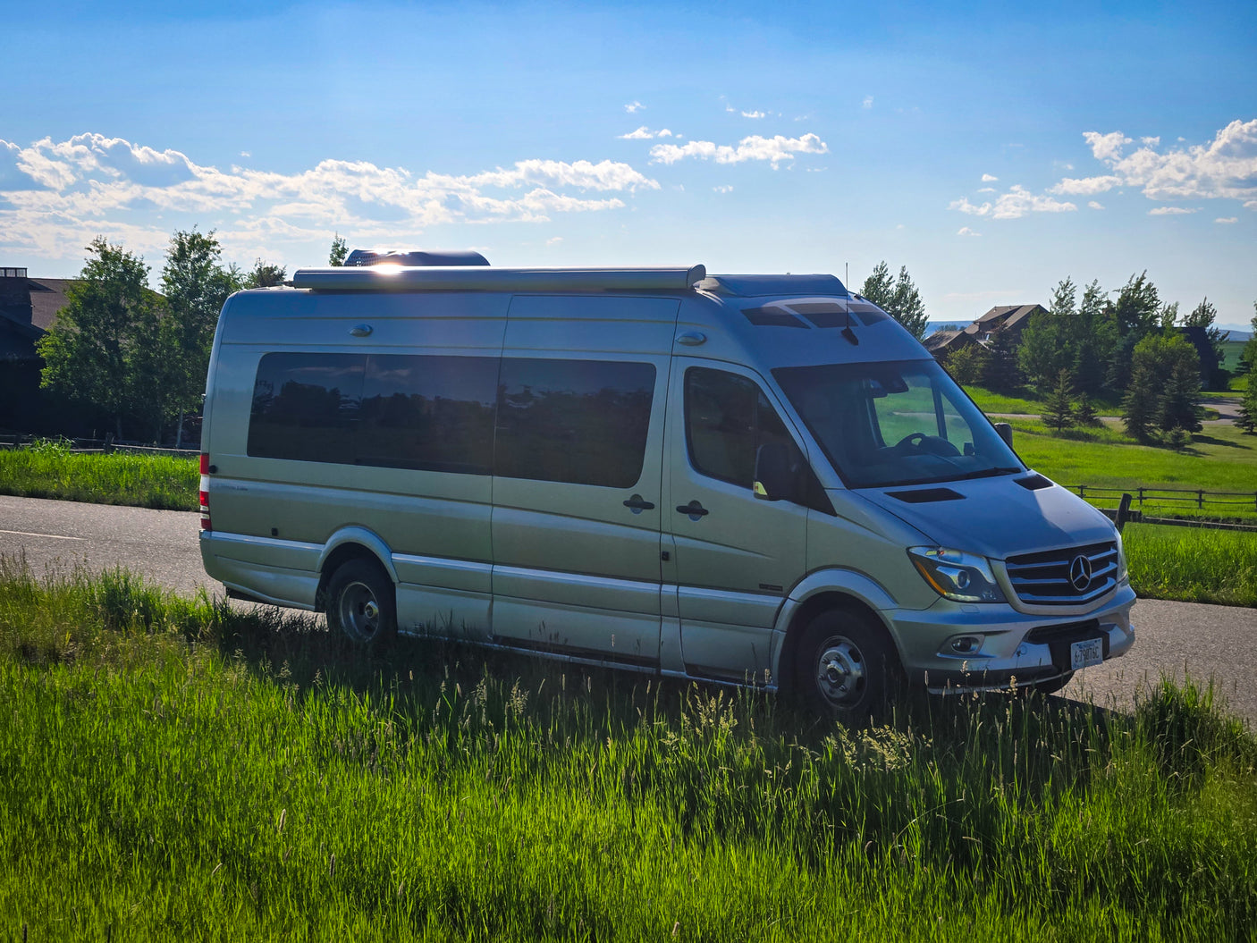 Silver Mercedes-Benz Sprinter camper van, an ideal RV rental, parked on a scenic road with lush grass & blue sky.