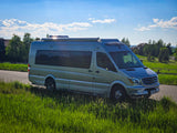 Silver Mercedes-Benz Sprinter camper van, an ideal RV rental, parked on a scenic road with lush grass & blue sky.