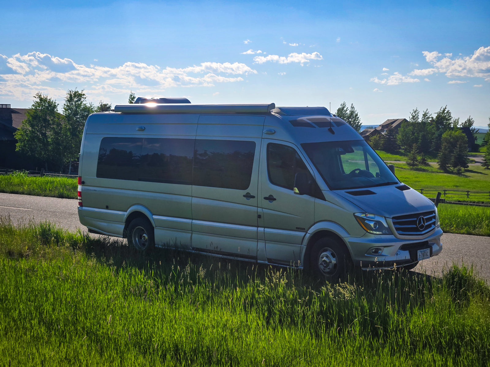 Silver Mercedes-Benz Sprinter camper van, an ideal RV rental, parked on a scenic road with lush grass & blue sky.