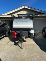 A sleek Bushwhacker travel trailer, ideal for rv rental, is parked in a sunny driveway near a house. Ready for trips!