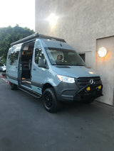 Custom grey Mercedes Sprinter RV rental. Child in driver's seat. Features off-road tires, light bar, and open side door.