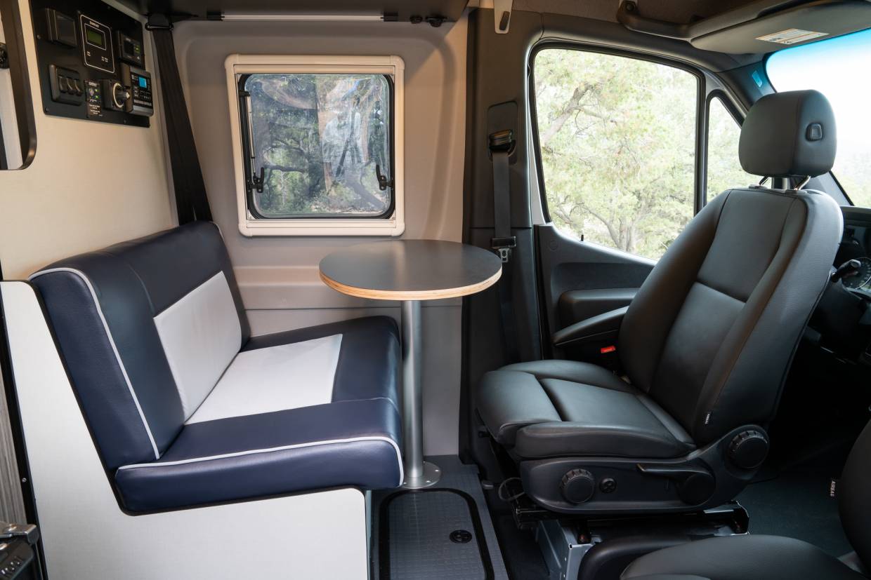 Inside this RV rental: a navy and white dinette with a round table, window, control panel, and black passenger seat.