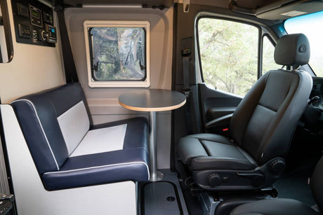 Inside this RV rental: a navy and white dinette with a round table, window, control panel, and black passenger seat.