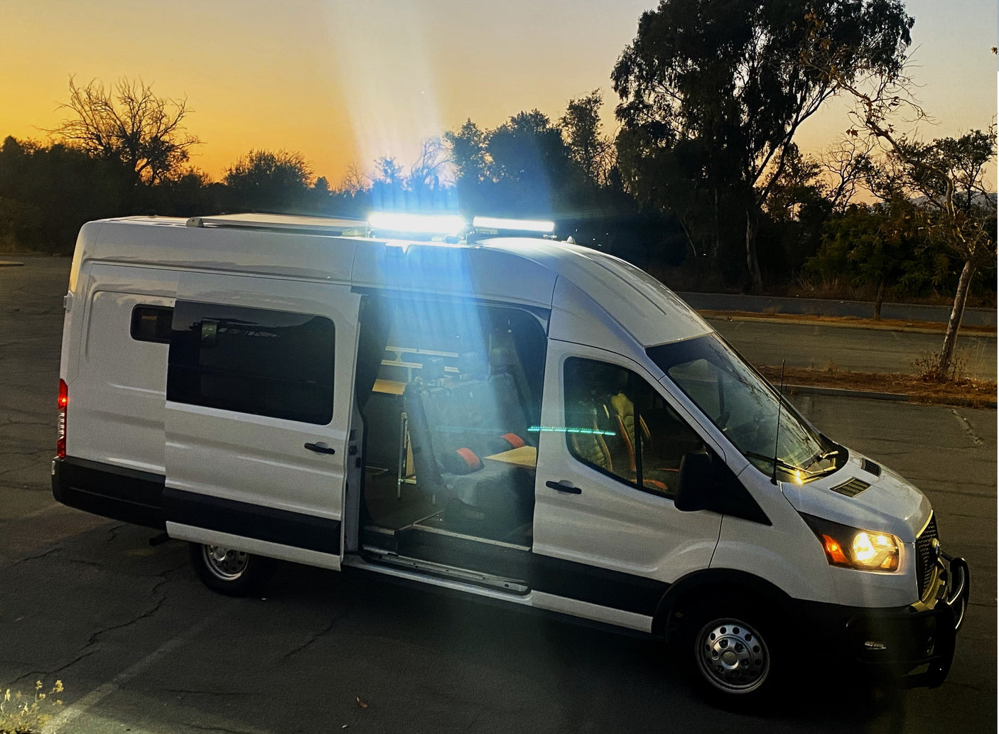 Brightly lit white RV rental camper van at sunset, doors open revealing interior setup. Perfect for your next road trip adventure.