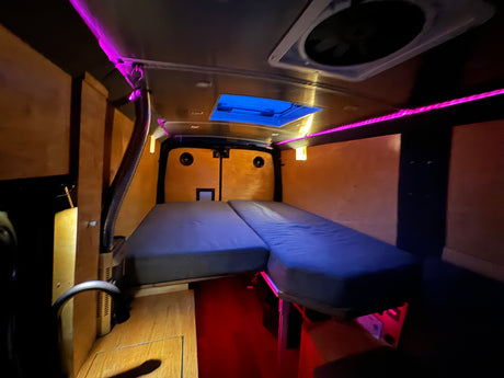Interior of an RV rental with a blue L-shaped bed, vibrant purple LED ceiling lights, and warm wooden paneling, creating a cozy sleeping area.