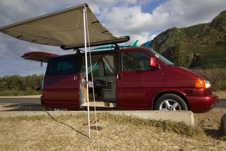 Red RV rental campervan with awning extended, interior visible through open doors, surfboards on roof, parked by green mountains.