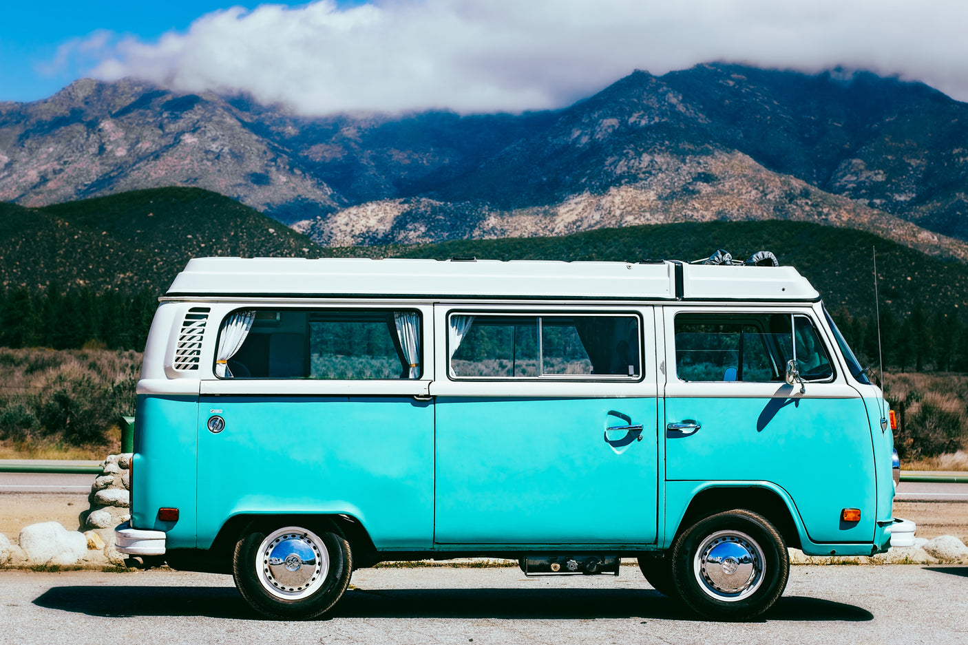 Vintage turquoise and white VW camper van, an ideal RV rental, parked roadside with mountains under a blue sky.