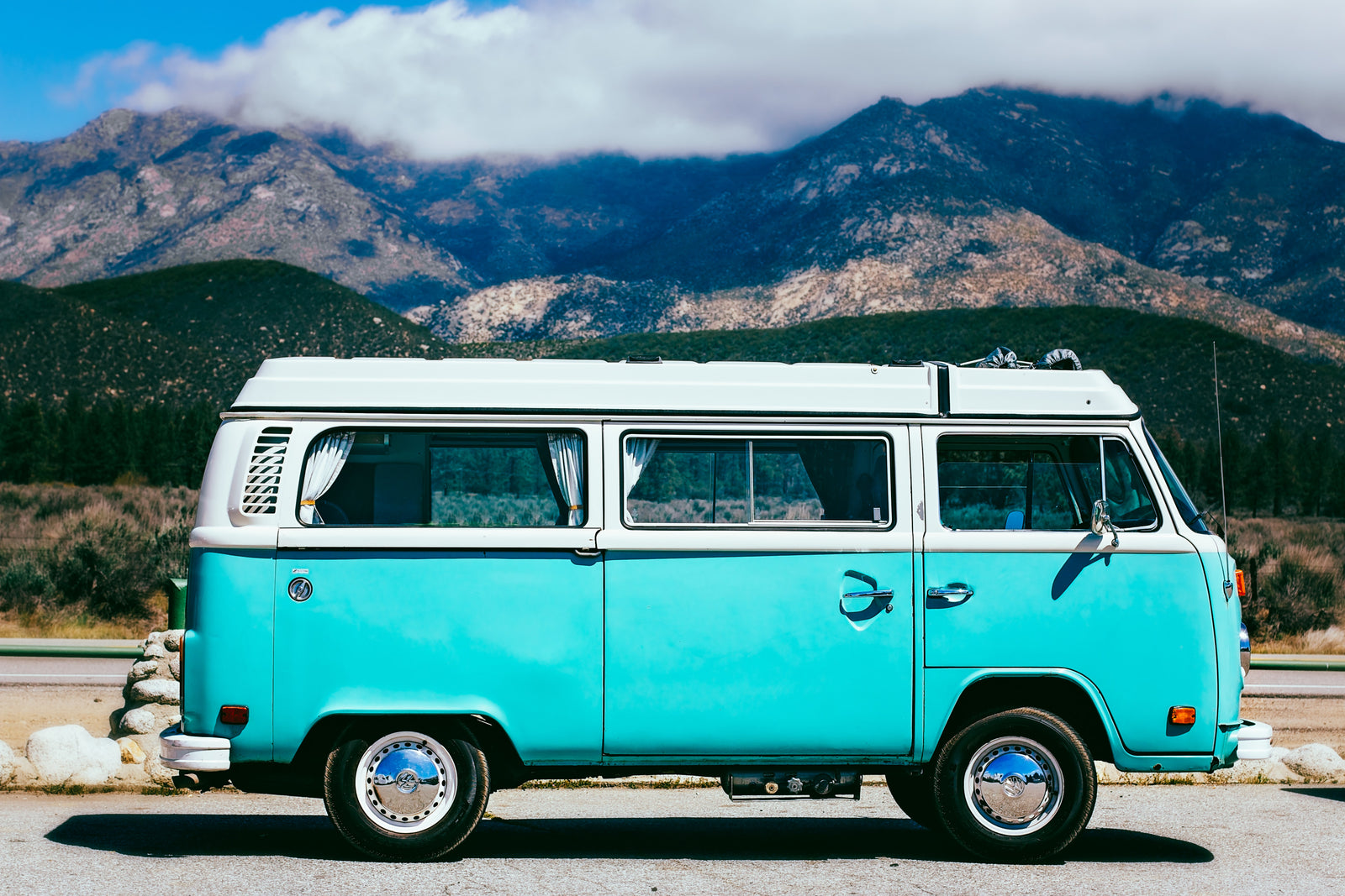 Vintage turquoise and white VW camper van, an ideal RV rental, parked roadside with mountains under a blue sky.