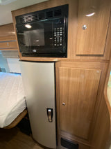 Compact kitchen area in an RV rental, featuring a built-in black microwave, silver refrigerator, and light wood cabinetry.
