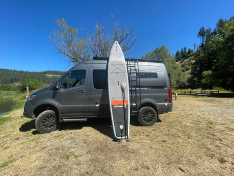 Experience van life! Gray camper van and paddleboard by water.
