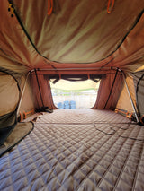 View inside a cozy RV rental rooftop tent: a quilted mattress on the floor and windows showing a backyard fence.