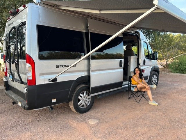 Relaxing next to a silver Sequence RV rental. A woman sits by its open door and extended awning, enjoying the outdoors.