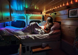 Woman relaxes in a cozy bed within an rv rental, illuminated by colorful string lights in the van's wood-paneled interior.