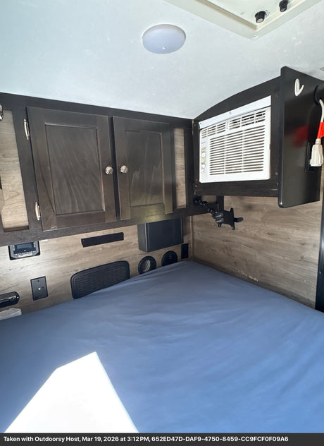 RV rental bedroom area showing a bed, dark wood overhead cabinets, and a wall-mounted AC unit, with wood-grain walls.