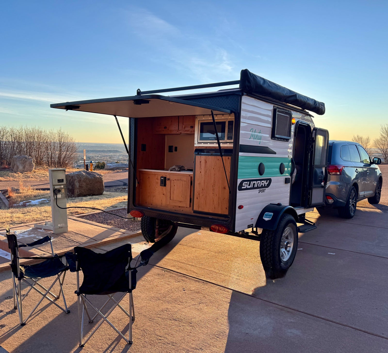 Sunray Sport RV rental: a teardrop camper with open kitchenette and power hookup, hitched to an SUV at a scenic campsite.