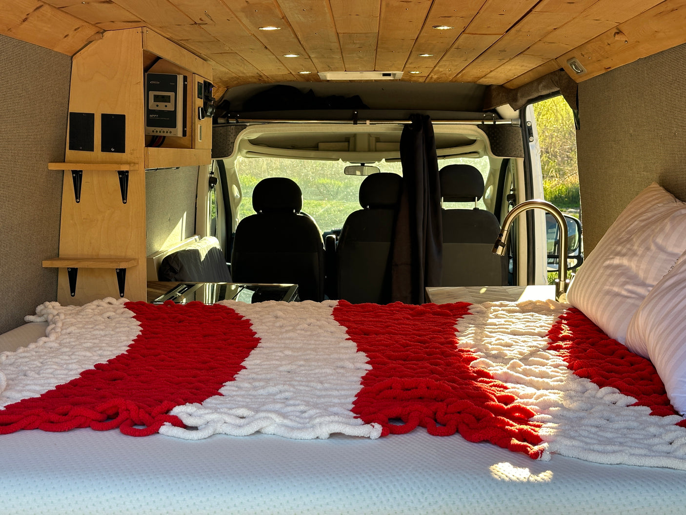Cozy RV rental interior: A bed with a red & white blanket, wooden shelves, electrical panel, and sink, all inside a camper van.