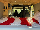 Cozy RV rental interior: A bed with a red & white blanket, wooden shelves, electrical panel, and sink, all inside a camper van.