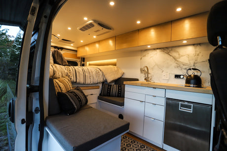 Bright RV rental interior: bed, cozy seating, and a sleek kitchen with sink, kettle, and fridge.