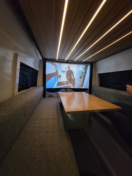 An RV rental's cozy dinette features a large projector screen displaying a van tour, illuminated by sleek ceiling LEDs.