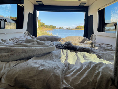 Cozy RV rental bed facing a stunning calm bay with trees. Soft blankets and pillows invite relaxation.