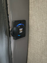 This RV rental features a dual USB charging port with a blue LED, mounted on a gray panel next to a textured wall.