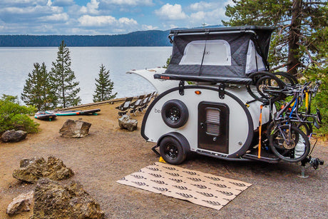 An RV rental teardrop trailer with a rooftop tent and bikes is parked at a scenic lake campsite with paddleboards.