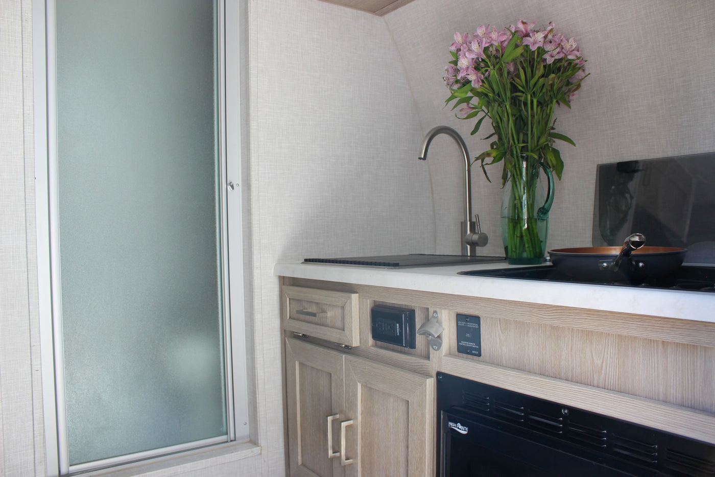 RV rental interior: a compact kitchen with sink, stove, and cabinet details, adjacent to a frosted shower. — photo 12