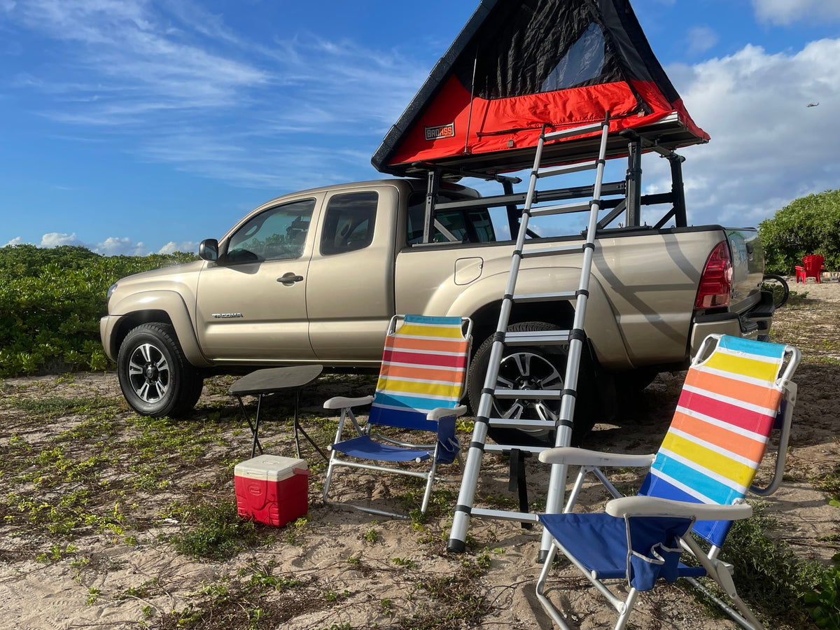 A tan pickup truck with a red rooftop tent and ladder, set up as an rv rental for camping. Beach chairs and cooler on sandy ground. — photo 18