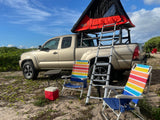 A tan pickup truck with a red rooftop tent and ladder, set up as an rv rental for camping. Beach chairs and cooler on sandy ground.